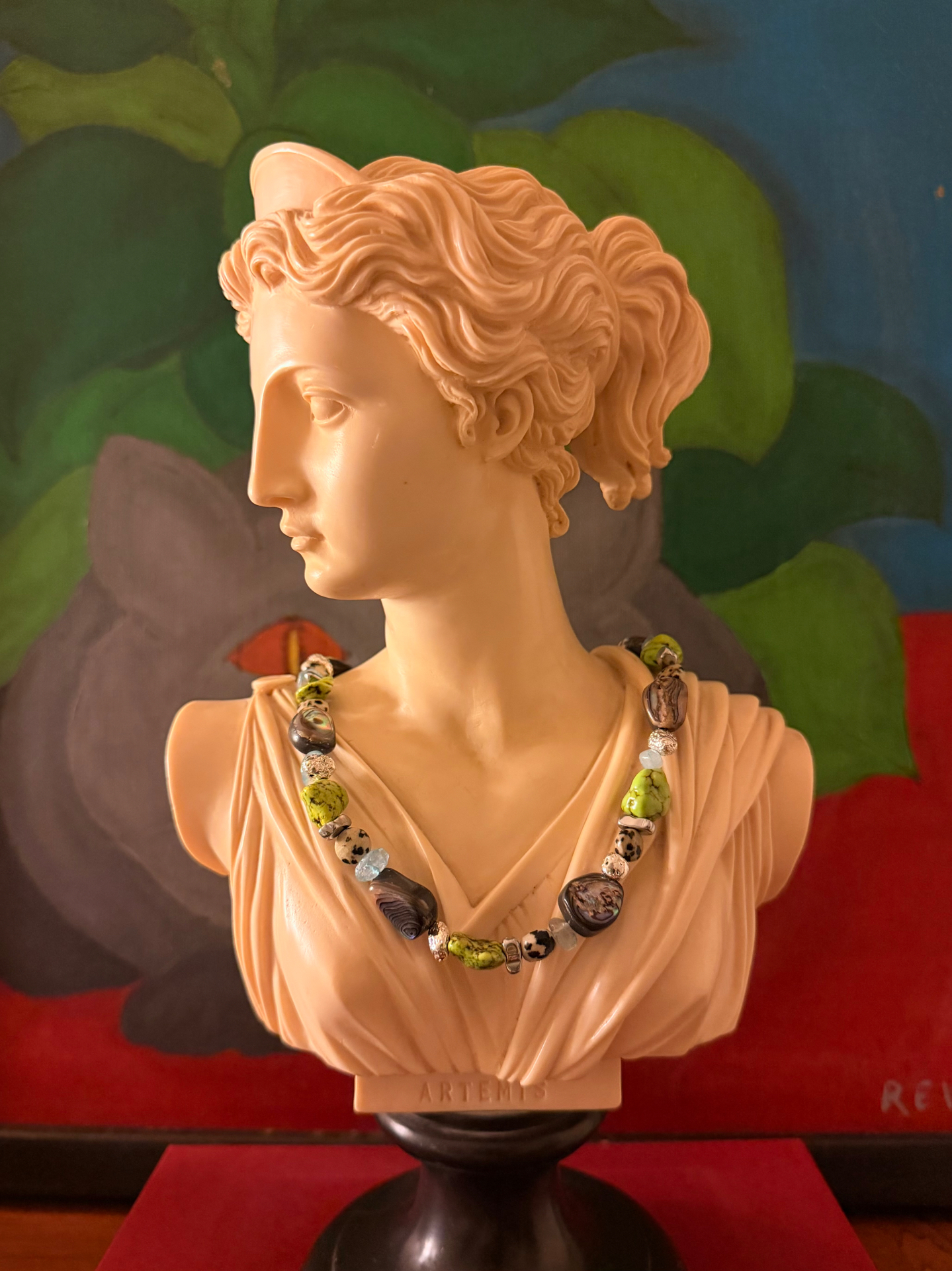 An Artemis bust wearing a hand-strung necklace of green turquoise, agate, and silver beads against a painted backdrop